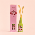 Cherry Soda reed diffuser in a green bottle with pink gift box and natural reeds on a soft peach background.