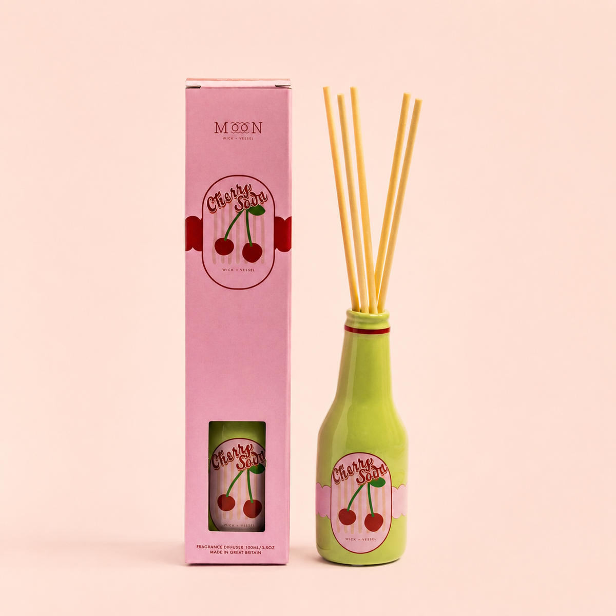 Cherry Soda reed diffuser in a green bottle with pink gift box and natural reeds on a soft peach background.