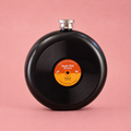 Round black hip flask designed to look like a vinyl record with grooved detailing, orange centre label and silver screw top on a pink background.