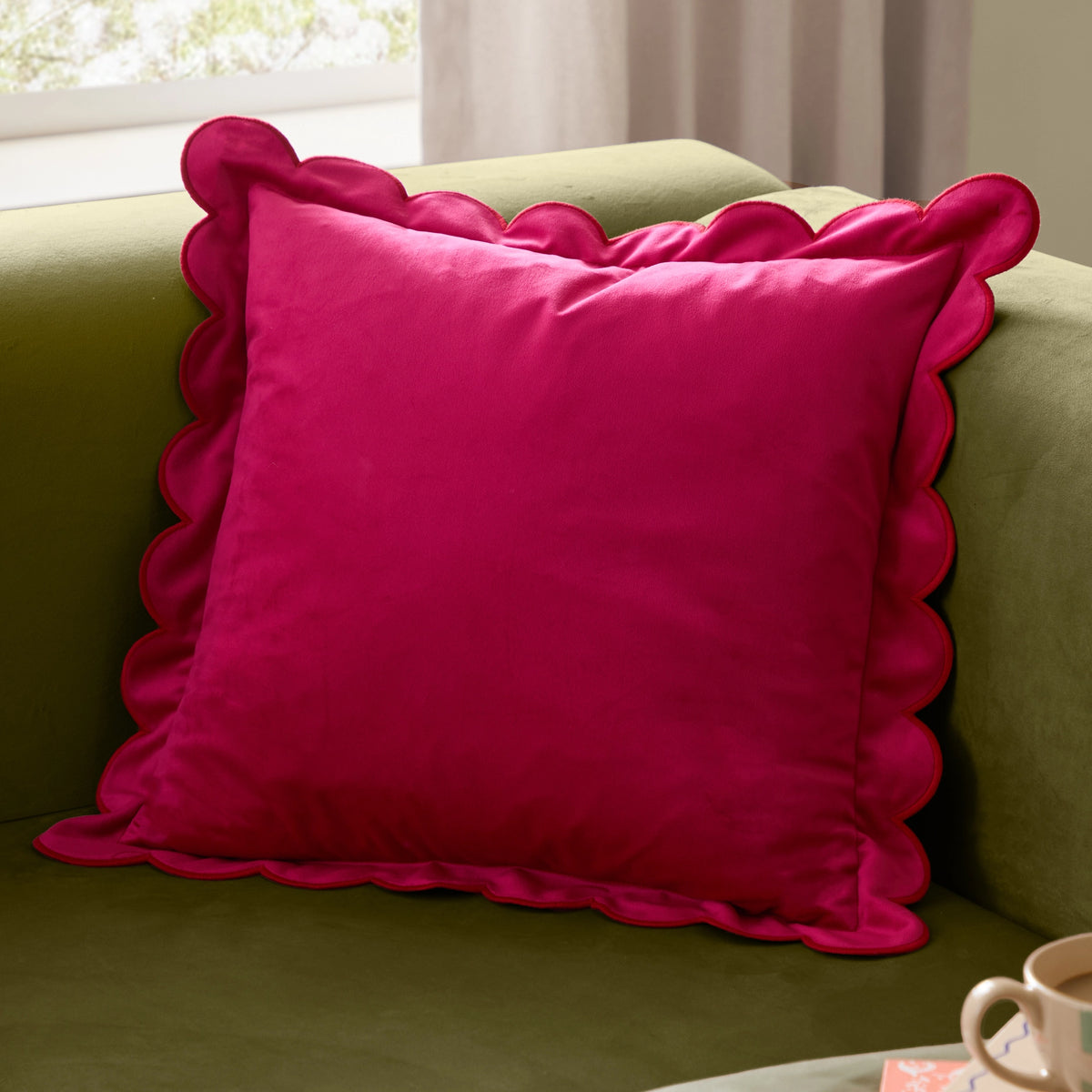 Hot pink velvet cushion with playful scalloped edging styled on an olive green sofa.
