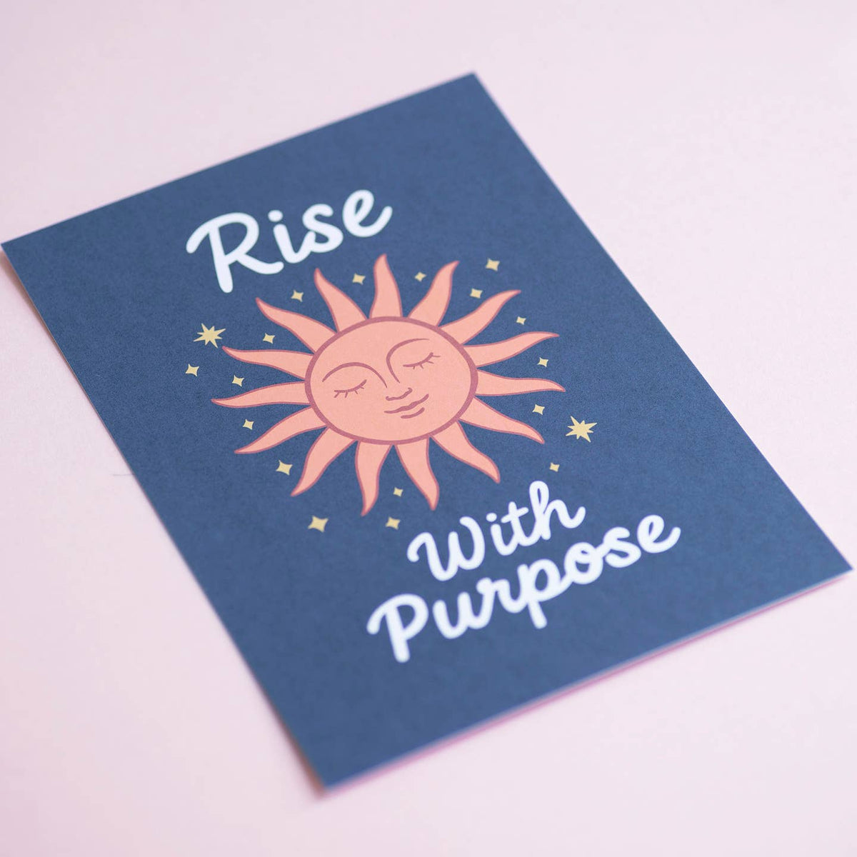 Colourful Rise With Purpose art print featuring a peaceful peach sun and golden stars, photographed against a soft pink background.