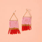 Pair of pink, red and gold beaded fringe earrings with triangular gold tone frames on a peach background.