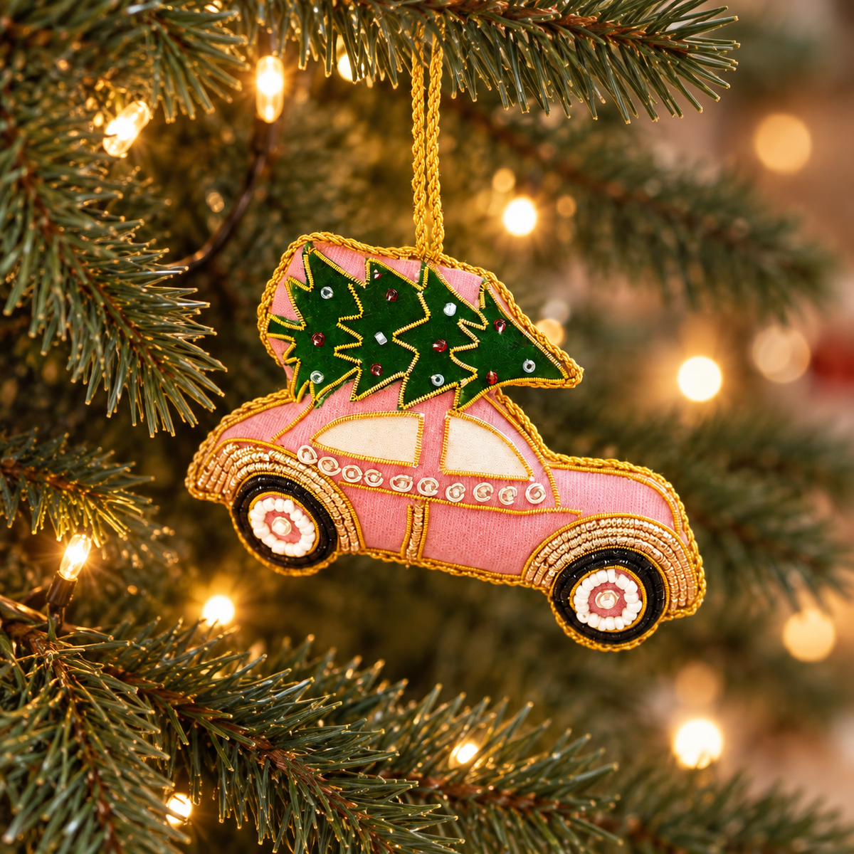Pink car Christmas bauble carrying green Christmas trees with gold beading, hanging on a lit Christmas tree.