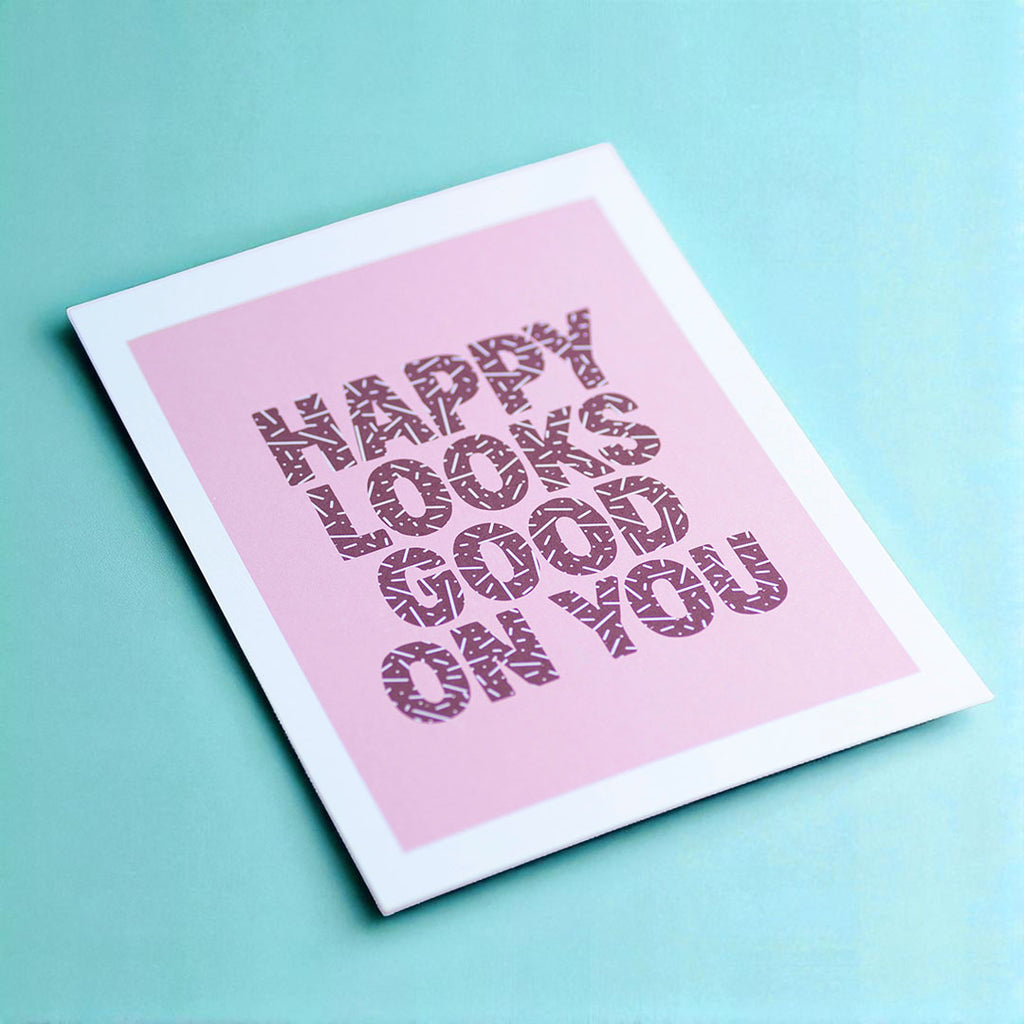 Happy Looks Good On You A5 print photographed at an angle, with patterned burgundy lettering and a soft pink background.