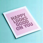 Happy Looks Good On You A5 print photographed at an angle, with patterned burgundy lettering and a soft pink background.