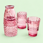 Pink fish stacking glasses partly assembled with two tumblers filled with drinks on a pale green background.