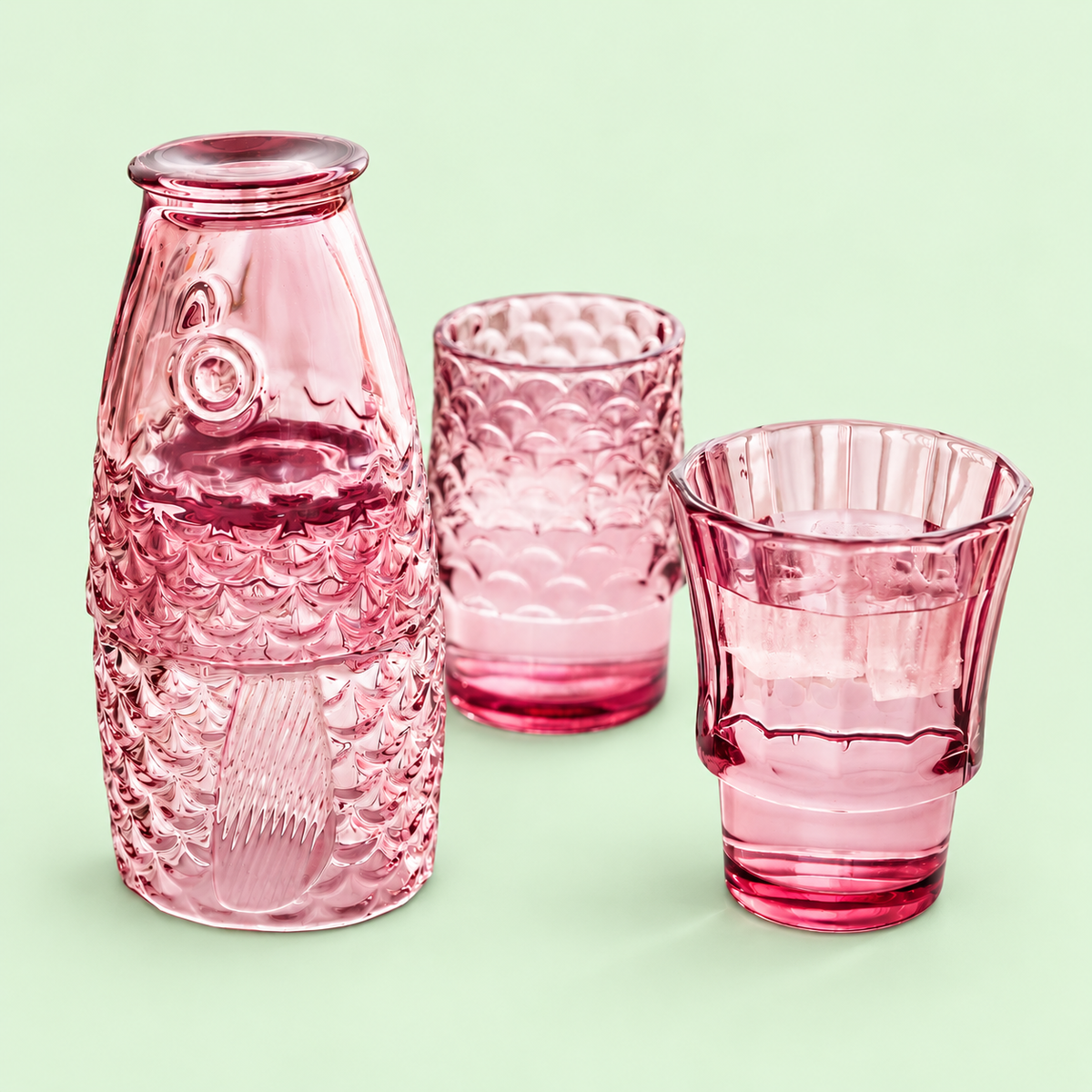 Pink fish stacking glasses partly assembled with two tumblers filled with drinks on a pale green background.
