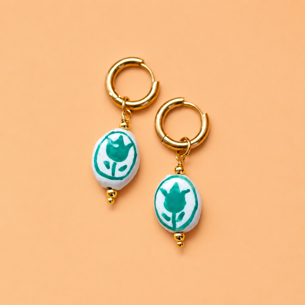 A pair of gold huggie hoop earrings with white oval charms featuring a green tulip design, displayed on a solid peach background.