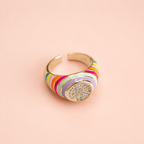 Adjustable gold tone ring with multicolour enamel stripes and sparkling round centre on a pale pink background.