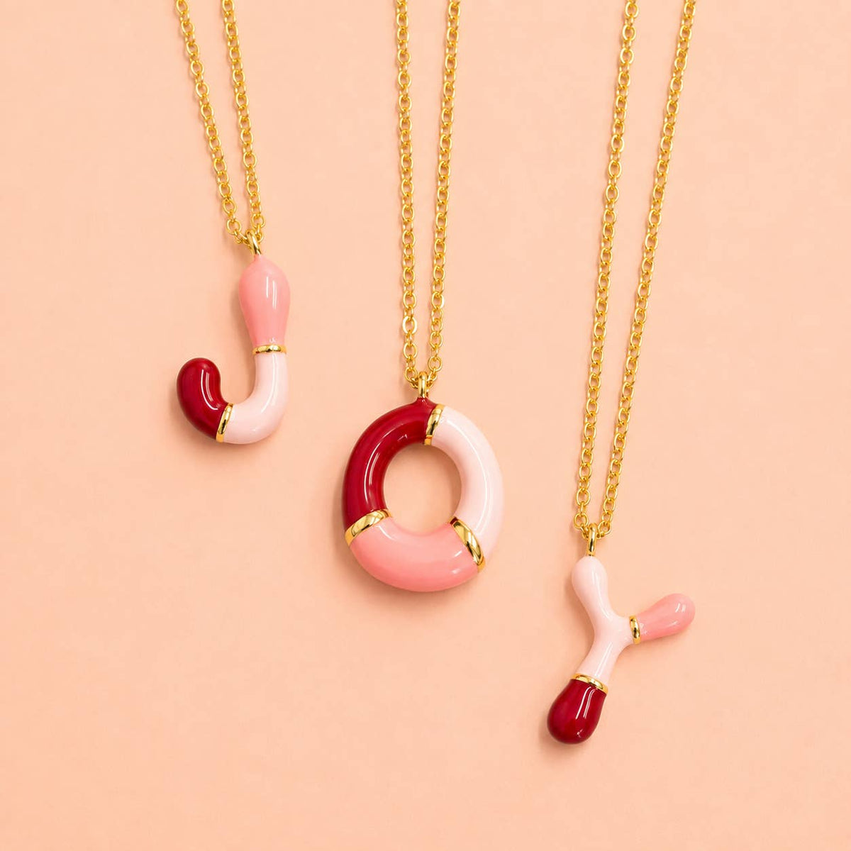 Pink and red enamel J, O and Y initial necklaces with gold-tone chains displayed on a soft pink background.