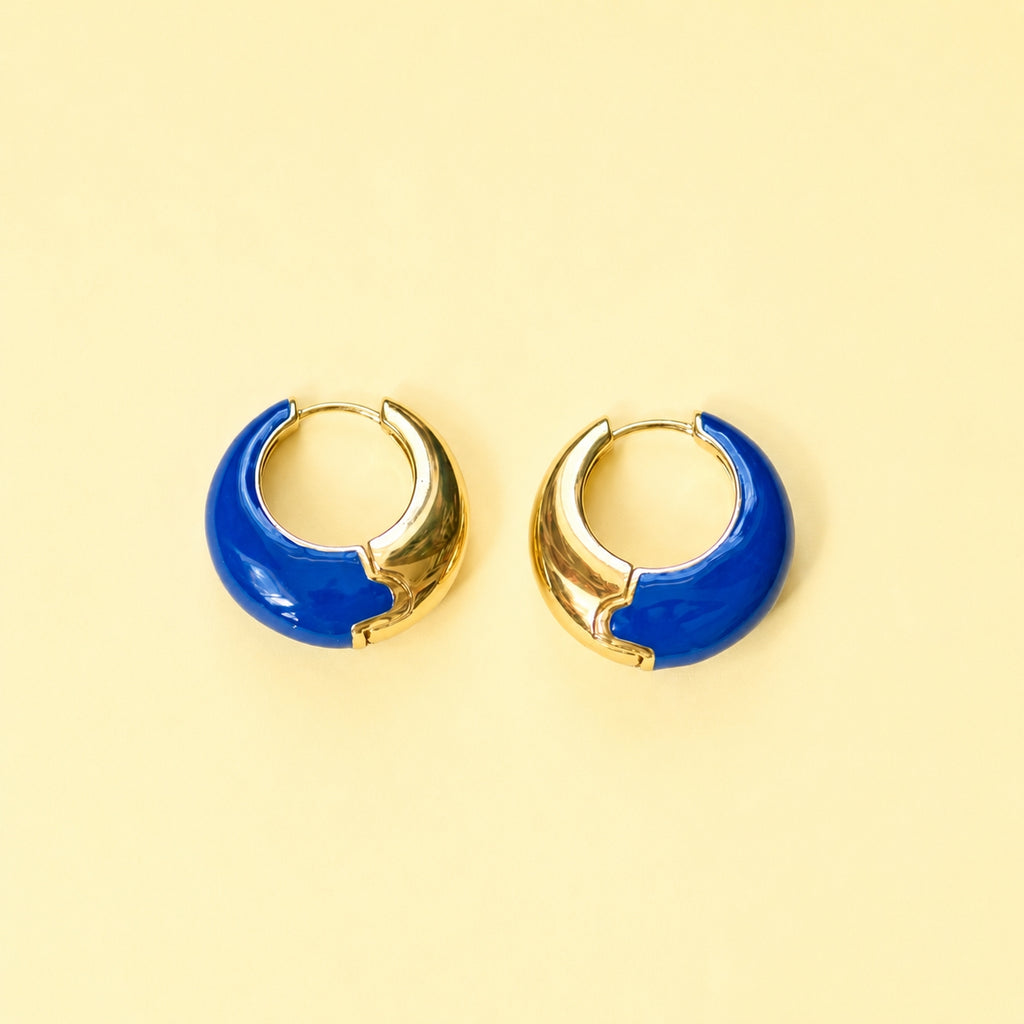 Cobalt blue enamel huggie hoop earrings with polished gold-tone detailing displayed on a yellow background.
