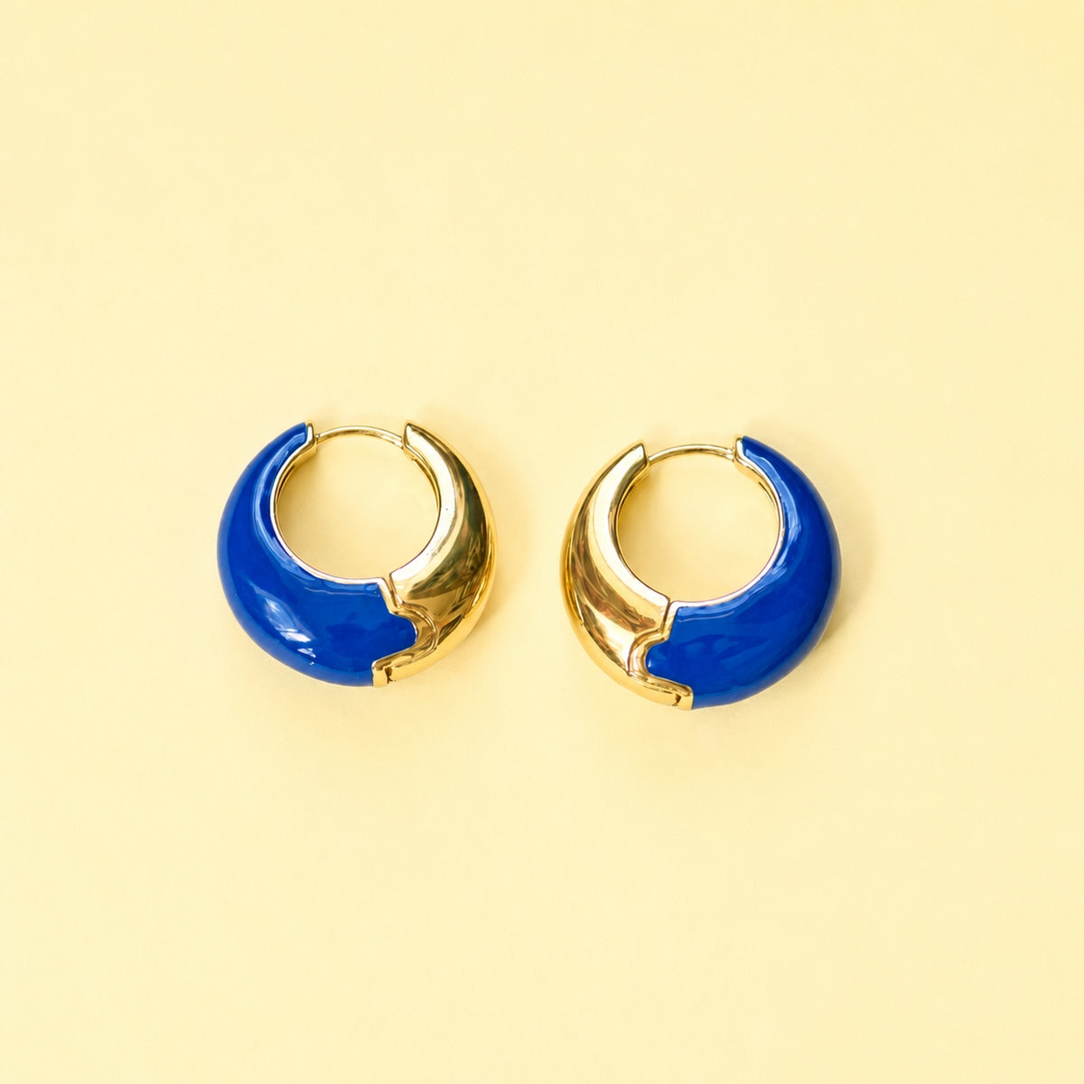 Cobalt blue enamel huggie hoop earrings with polished gold-tone detailing displayed on a yellow background.
