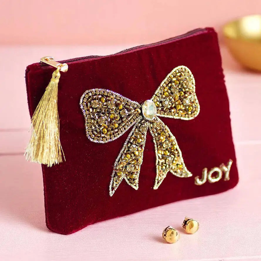 Red velvet pouch with sparkling gold beaded bow, ‘Joy’ lettering and gold tassel zip pull displayed on a pink background.