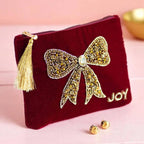 Red velvet pouch with sparkling gold beaded bow, ‘Joy’ lettering and gold tassel zip pull displayed on a pink background.