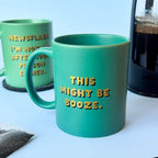 Green ceramic mug reading This Might Be Booze in gold and black lettering beside a coffee pot and another slogan mug.