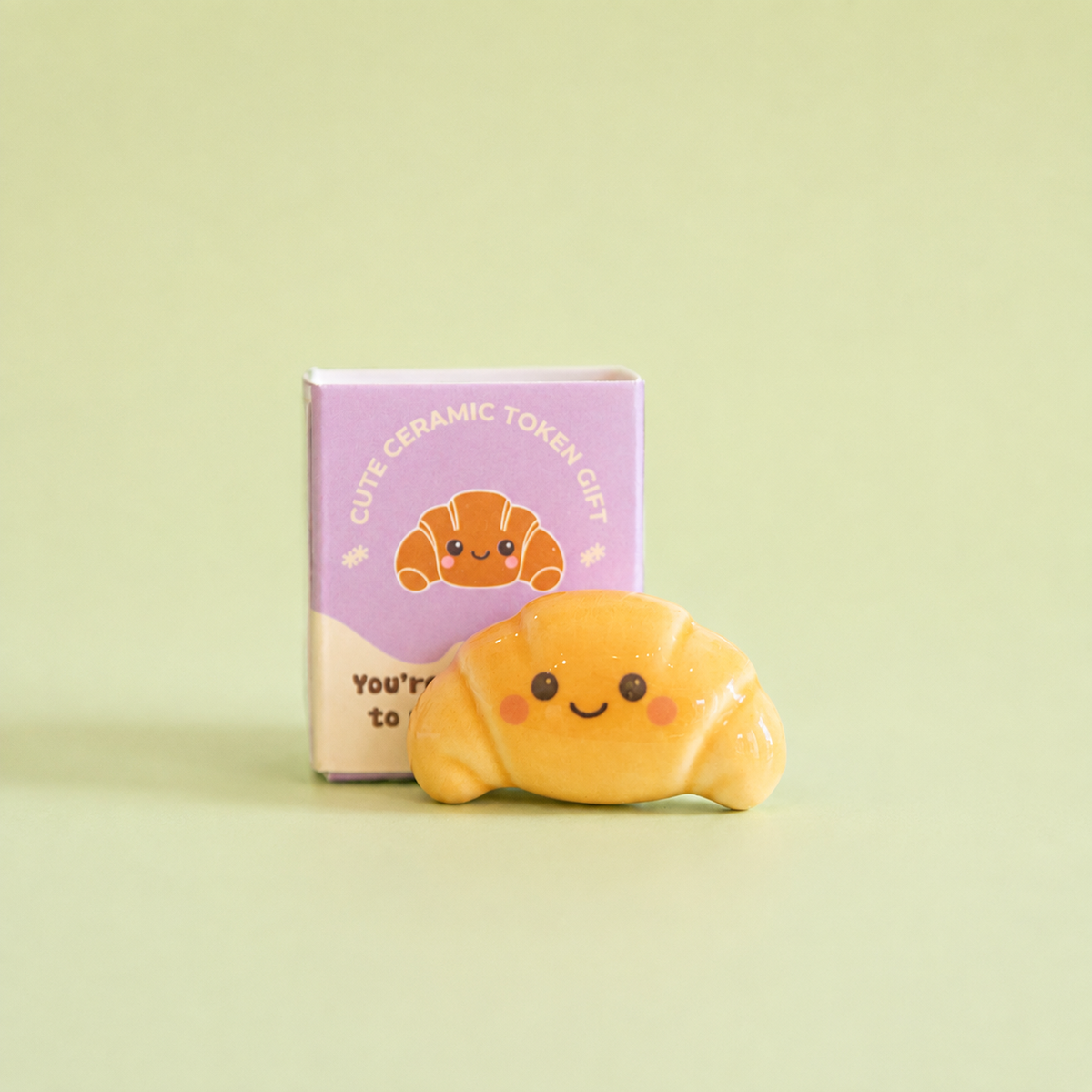 Cute smiling ceramic croissant token displayed in front of its purple illustrated gift box on a pale green background.