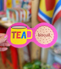 Bright pink double coaster with yellow tea mug, colourful TEA lettering and illustrated biscuit, held inside a colourful shop.