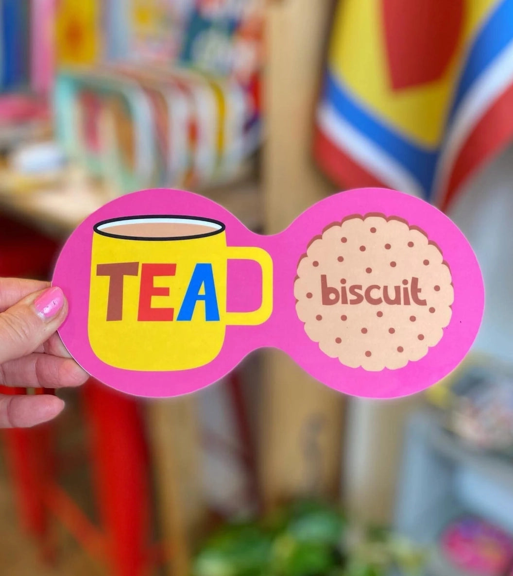 Bright pink double coaster with yellow tea mug, colourful TEA lettering and illustrated biscuit, held inside a colourful shop.