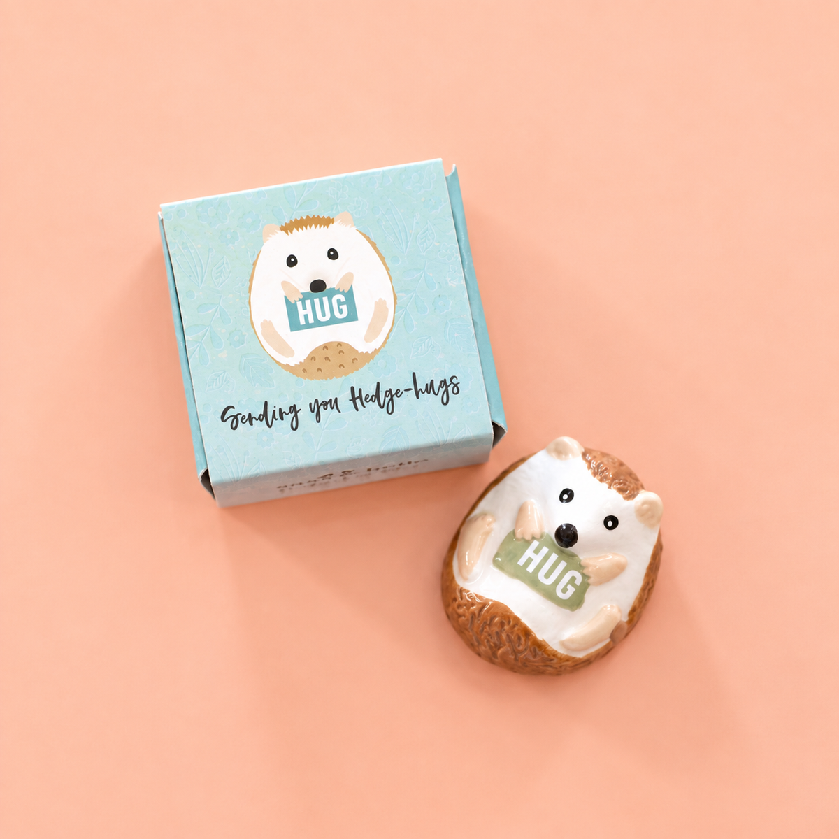 Ceramic hedgehog token holding a HUG sign beside its illustrated blue gift box on a peach pink background.