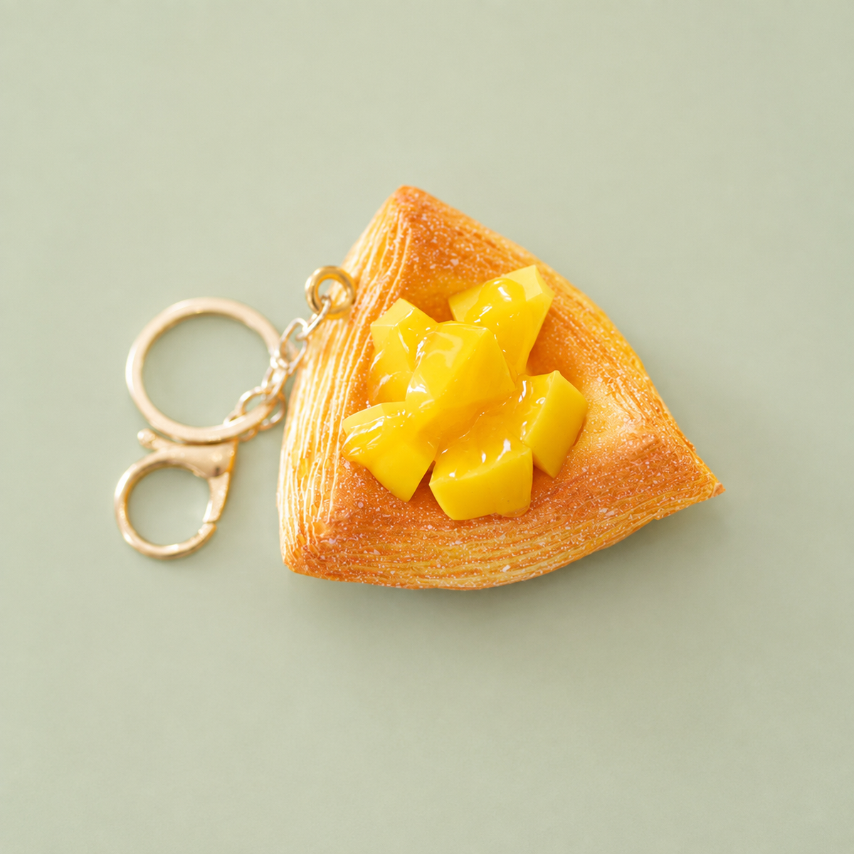Mango Danish pastry bag charm keyring with golden pastry layers, glossy yellow mango pieces and gold clip on a pale green background.