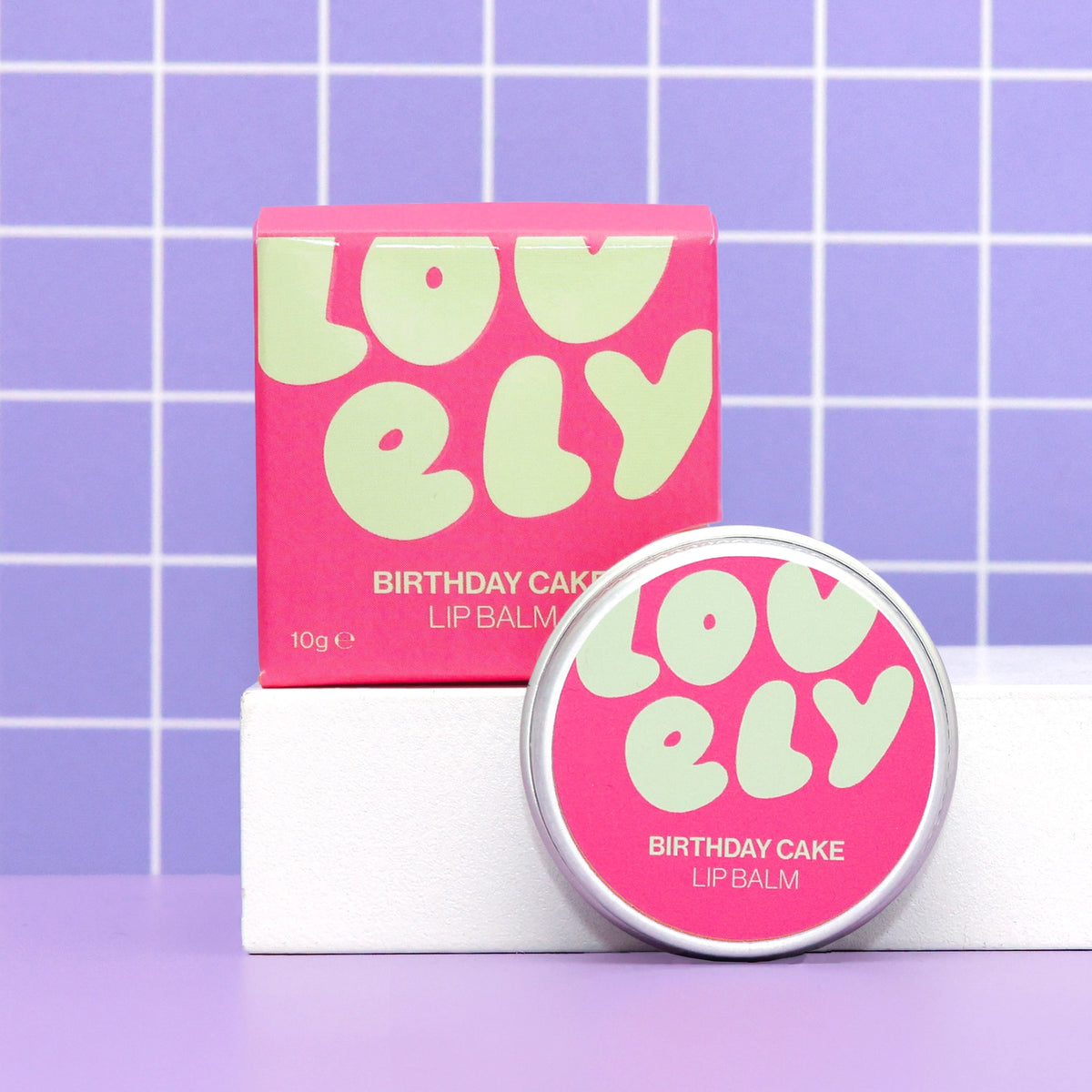Birthday Cake Lip Balm tin and colourful pink box displayed against a lilac tiled background.