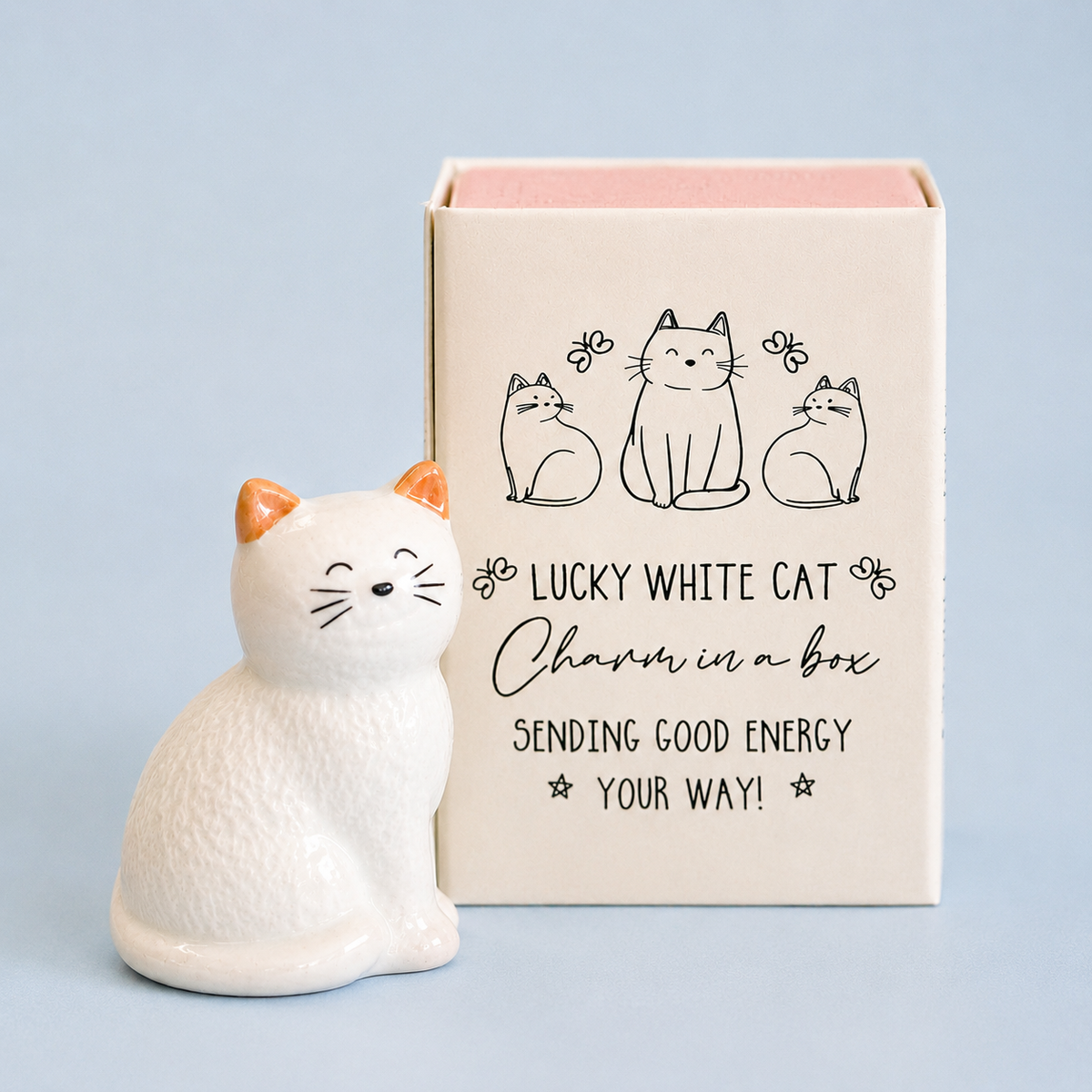 White ceramic lucky cat token with peach ears beside its illustrated gift box on a soft pale blue background.