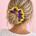 Yellow, grey and hot pink crochet scrunchie worn around a blonde messy bun against a pale pink background.