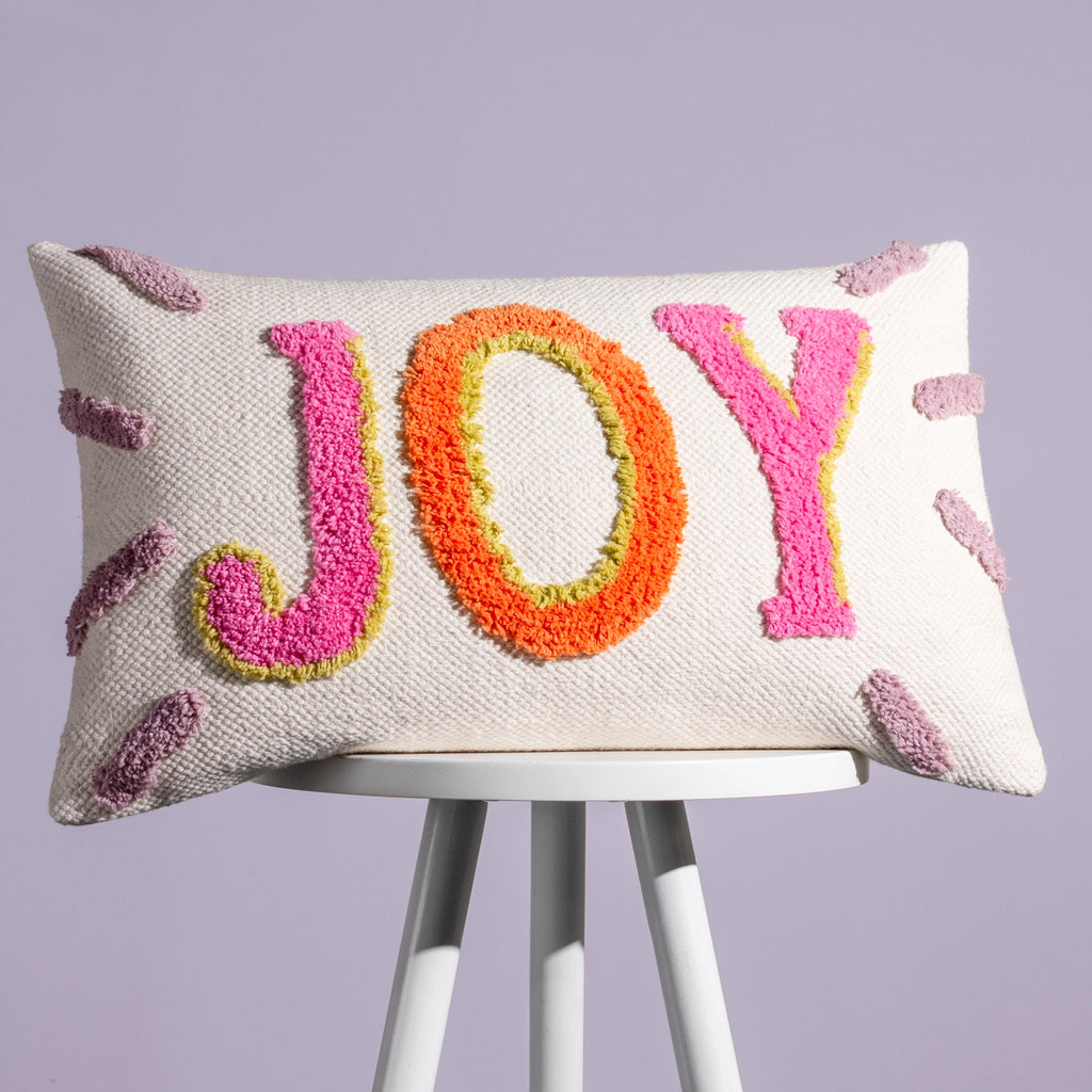 Cream JOY cushion with colourful tufted pink, orange and green lettering displayed on a stool.