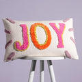Cream JOY cushion with colourful tufted pink, orange and green lettering displayed on a stool.