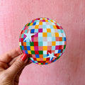 Colourful round disco ball coaster with multicoloured checkerboard design and white stars, held against a pink background.