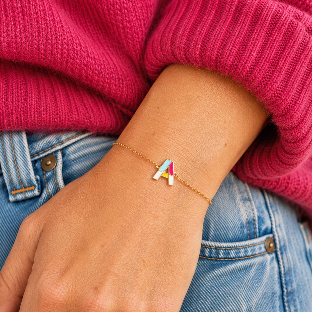 Colourful enamel letter A bracelet on a delicate gold-tone chain, styled with a bright pink jumper and denim.
