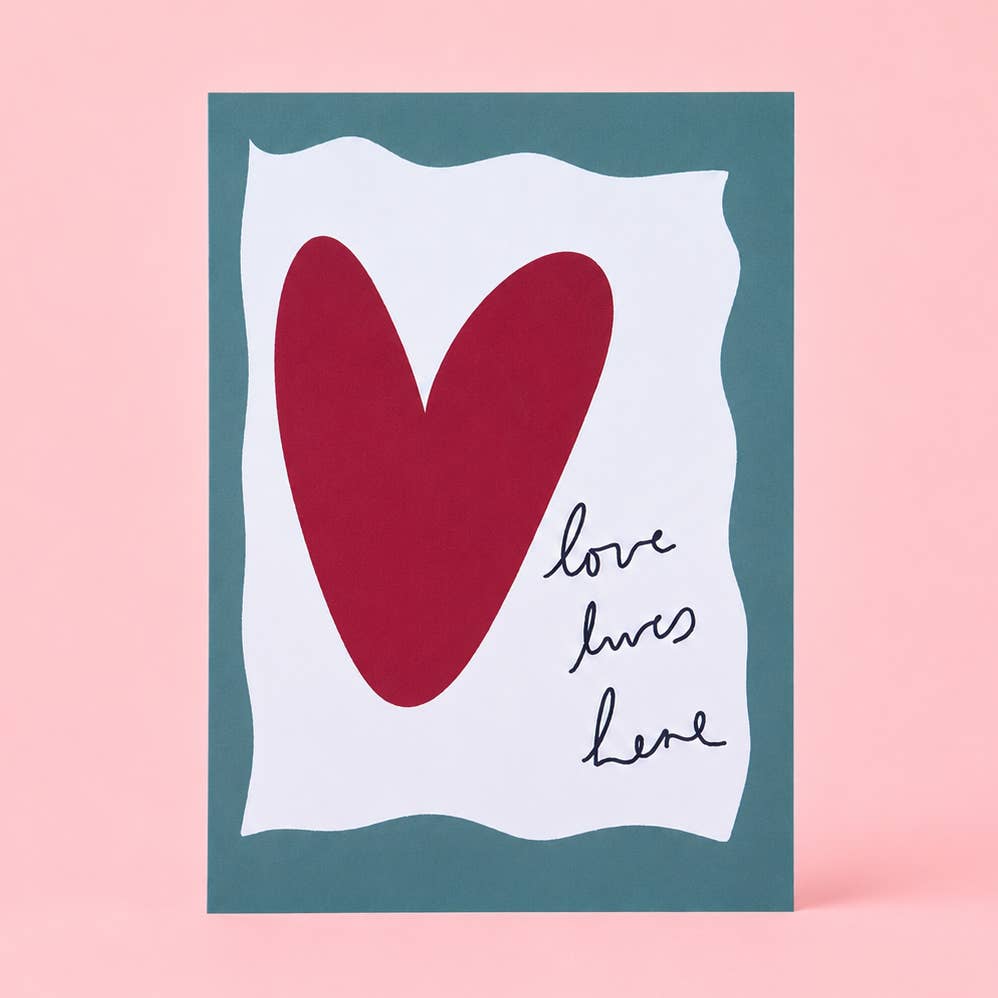 Heartwarming Love Lives Here A5 art print with a bold red heart, handwritten lettering and deep teal border on a soft pink background.