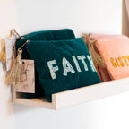 Teal velvet ‘FAITH’ pouches with beaded lettering and gold tassels displayed alongside a pink beaded pouch.