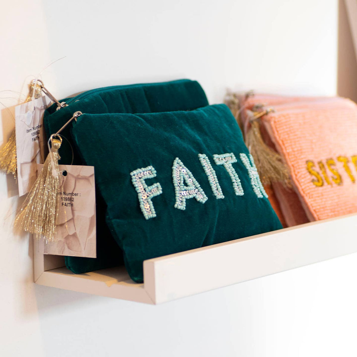 Teal velvet ‘FAITH’ pouches with beaded lettering and gold tassels displayed alongside a pink beaded pouch.
