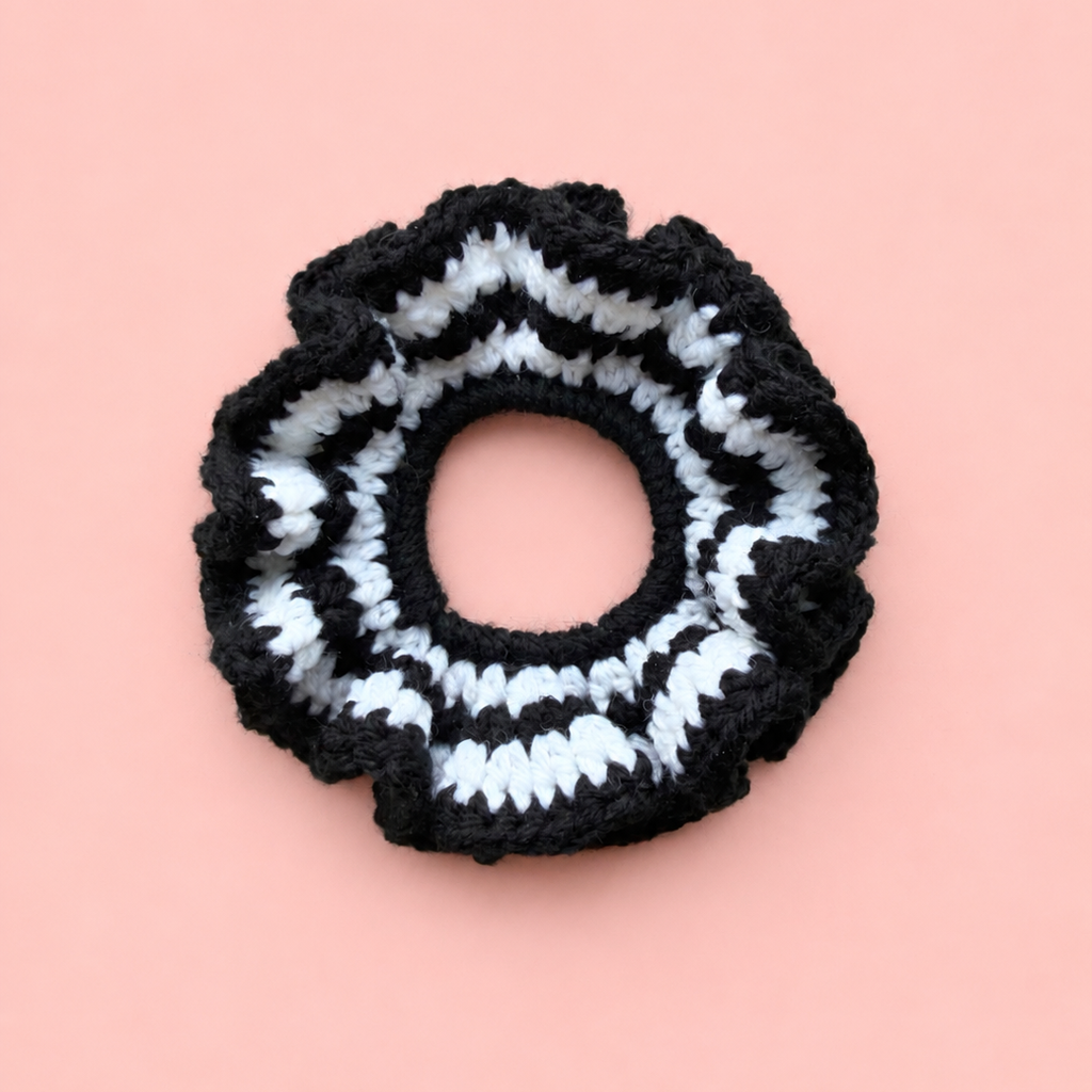 One of a kind handmade black and white striped crochet scrunchie with a chunky ruffled design on a soft pink background.