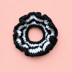 One of a kind handmade black and white striped crochet scrunchie with a chunky ruffled design on a soft pink background.