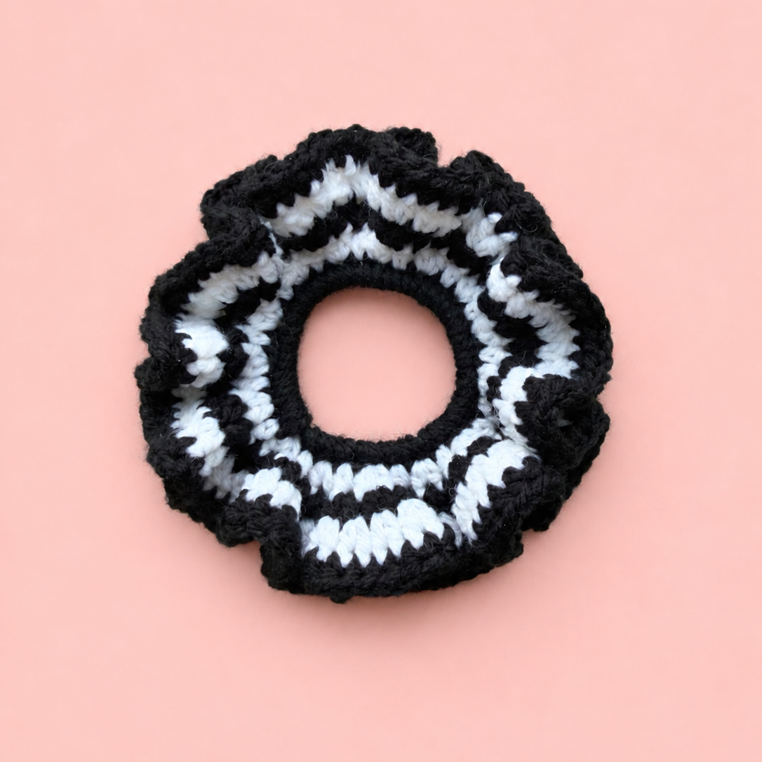 One of a kind handmade black and white striped crochet scrunchie with a chunky ruffled design on a soft pink background.