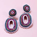 Pink and blue beaded oval statement earrings with layered seed bead detailing on a pale pink background.