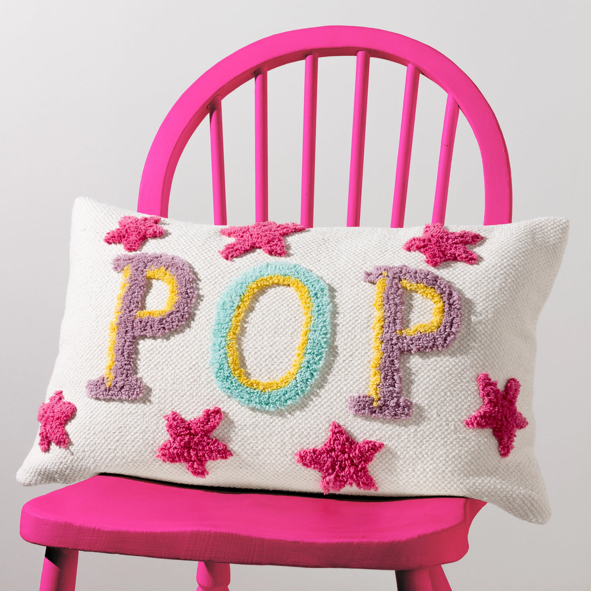 Cream rectangular POP cushion with colourful raised lettering and pink stars styled on a bright pink chair.