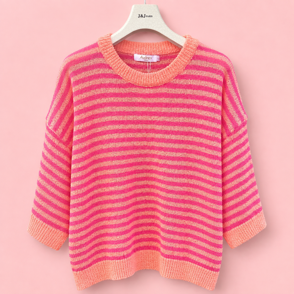 Coral and bright pink striped knitted jumper with three quarter sleeves on a hanger against a soft pink background.