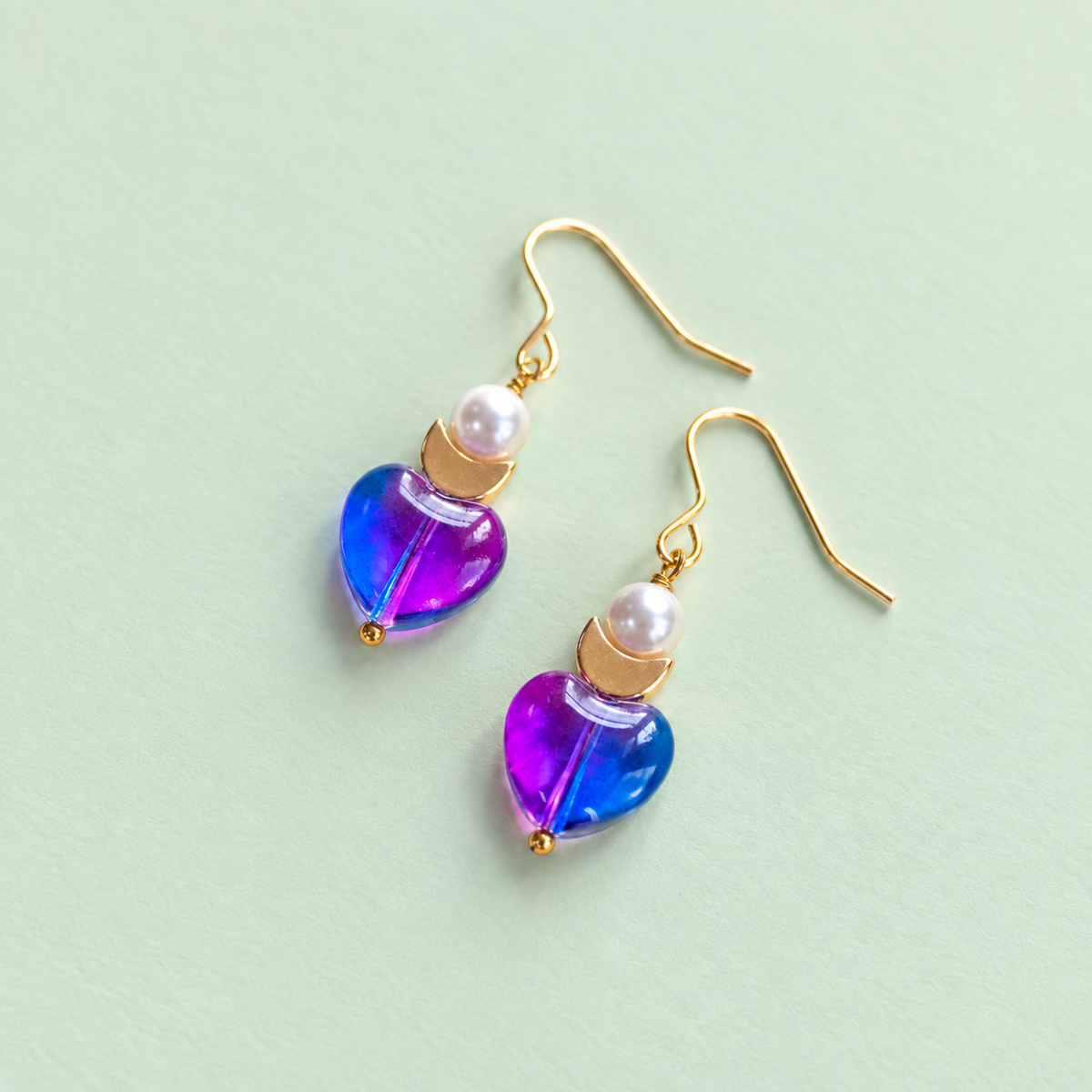 One of a kind gold plated drop earrings with pink and blue glass heart beads, pearl details and small gold crescent moons on a pale green background.