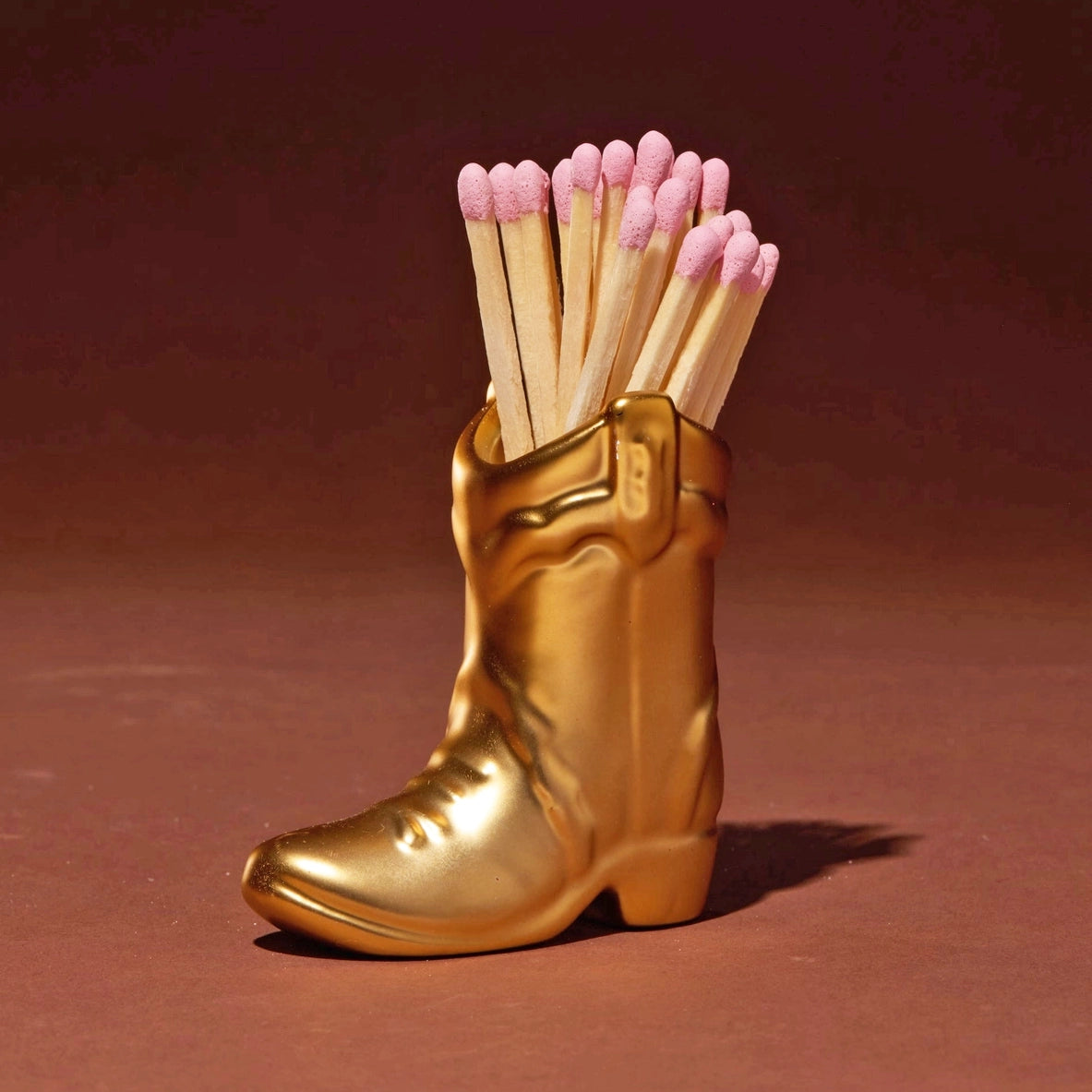 Gold ceramic cowboy boot match holder filled with pink tipped matches on a rich brown background.