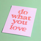 Pink Do What You Love A5 print with playful coral red and white heart lettering displayed on a fresh green background.