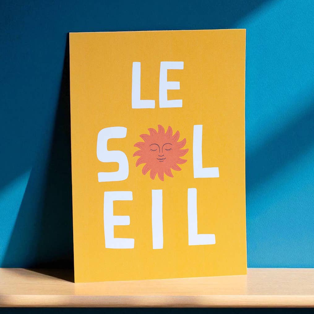 Bright yellow Le Soleil A5 art print with white lettering and a smiling coral sun displayed against a vivid blue wall.