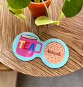 Aqua blue double coaster with bright pink tea mug, colourful TEA lettering and illustrated biscuit on a wooden table.