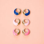 Three pairs of colourful enamel huggie hoop earrings in cobalt blue, pale pink and bright pink with gold-tone detailing.