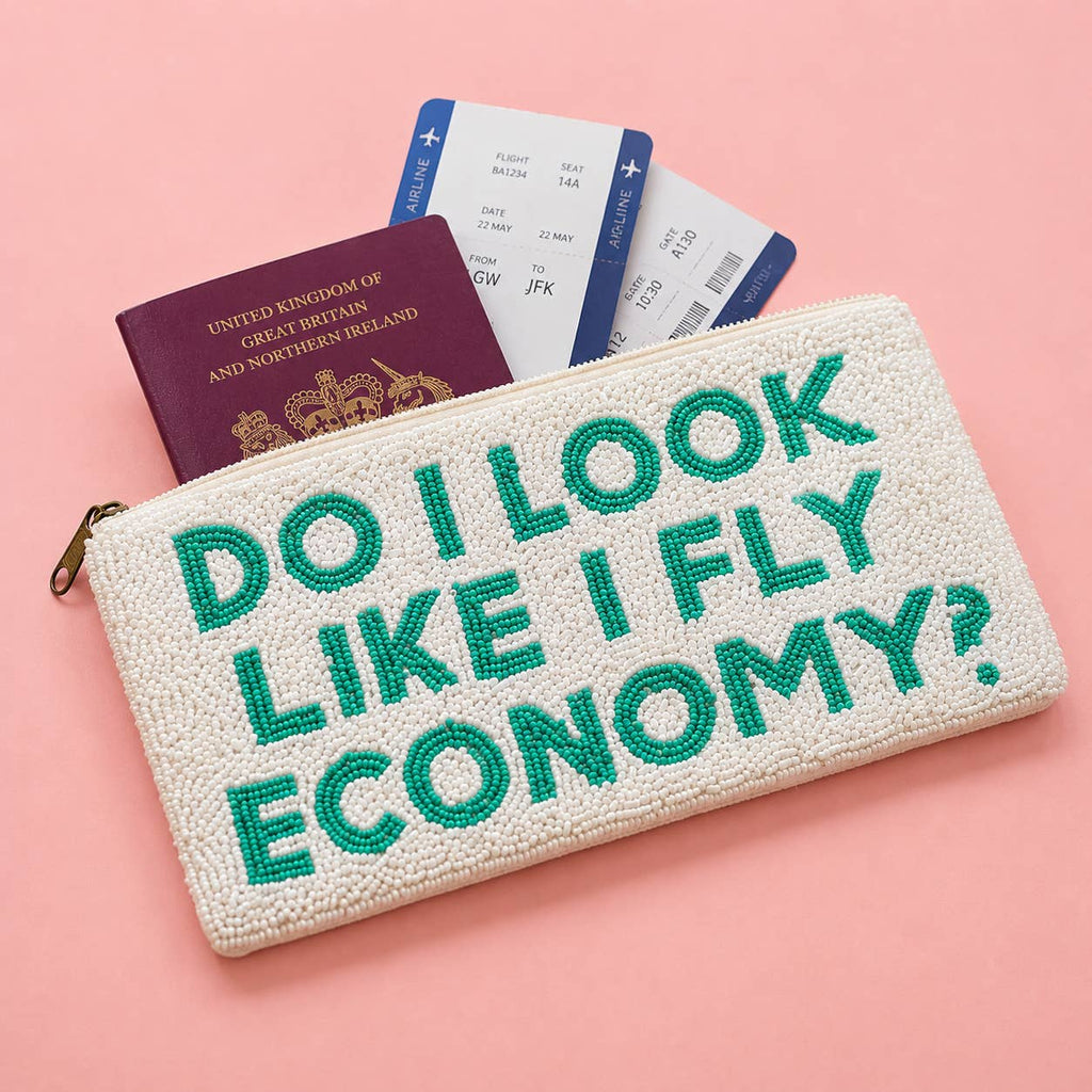 Cream beaded travel wallet with turquoise ‘Do I Look Like I Fly Economy?’ lettering beside a passport and boarding passes.