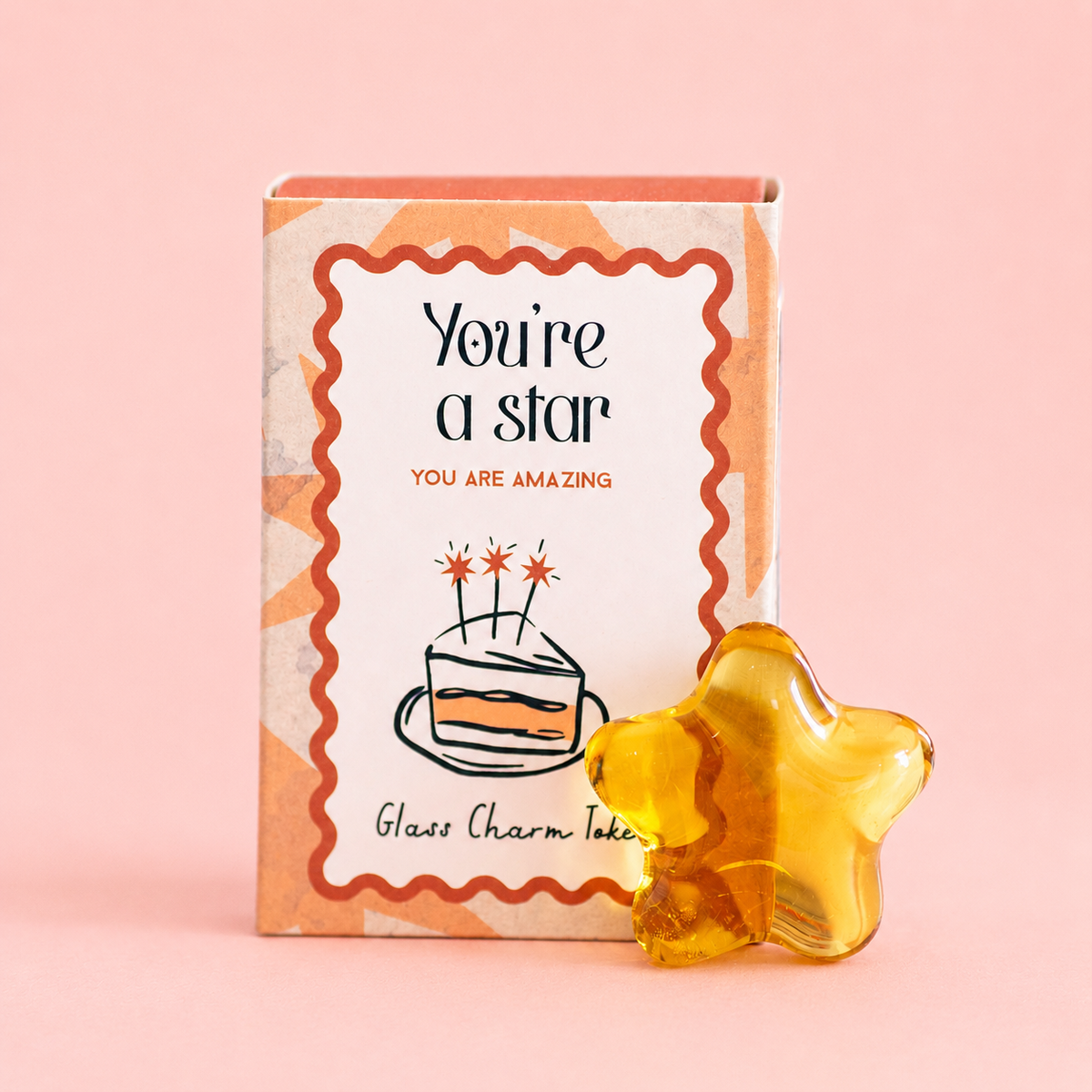 Golden glass star token beside its You’re A Star gift box with You Are Amazing message on a soft pink background.