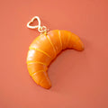 Realistic golden croissant bag charm keyring with pastry detailing and a gold heart shaped clip.
