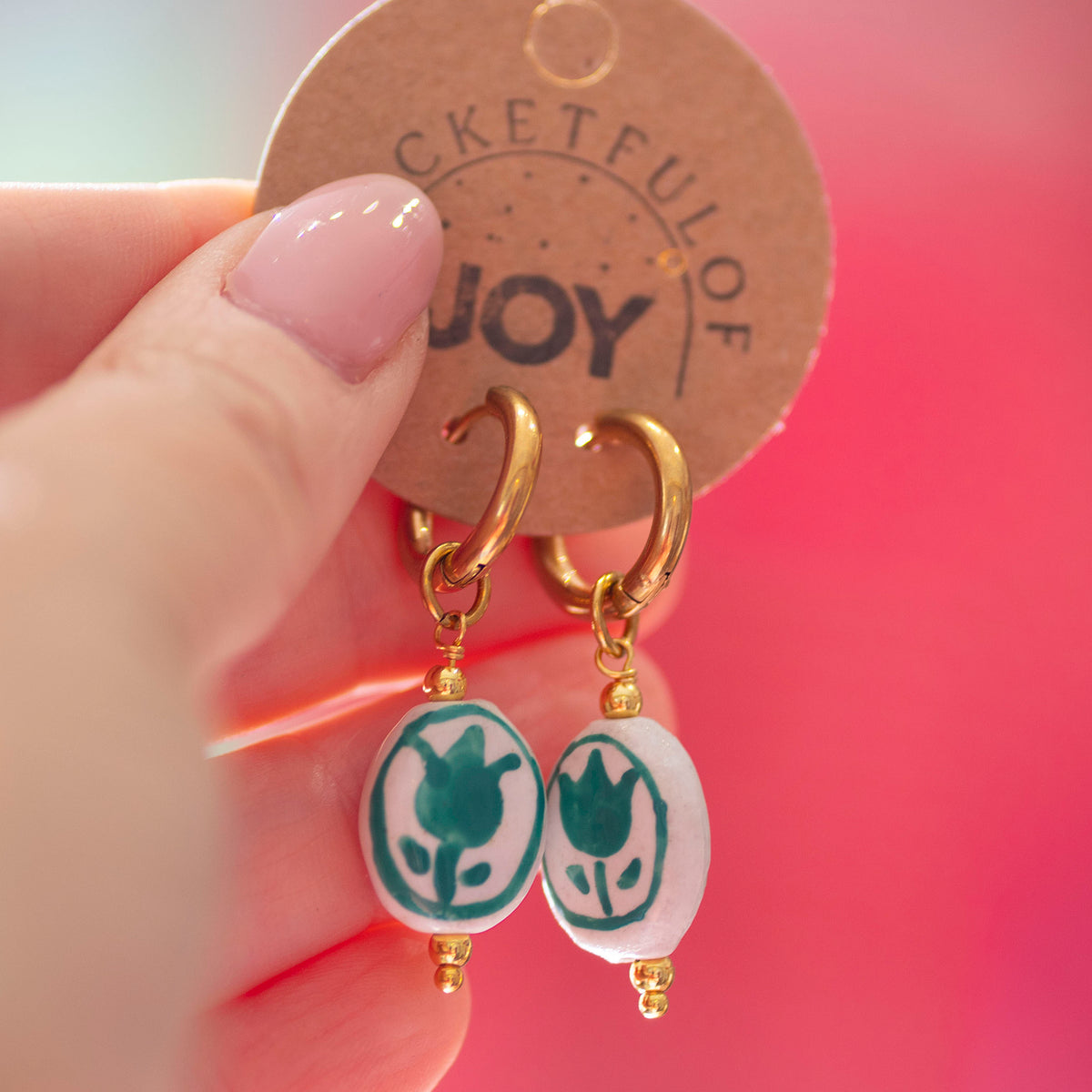 A close-up of a hand holding a round brown paper tag that displays a pair of gold hoop earrings with white oval beads featuring a teal tulip design.